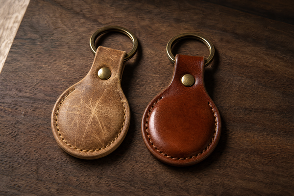 Two leather AirTag keychains side by side on walnut, the left in pale waxed Crazy Horse leather covered in fine scratch lines, the right in chestnut vegetable tanned leather with a smooth burnished sheen