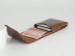 A brown leather card holder wallet shown open, containing a pink banknote and credit cards, on a light gray background. 