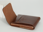 A brown leather card holder wallet with an open flap, showing a card slot and white stitching, photographed on a light gray background. 