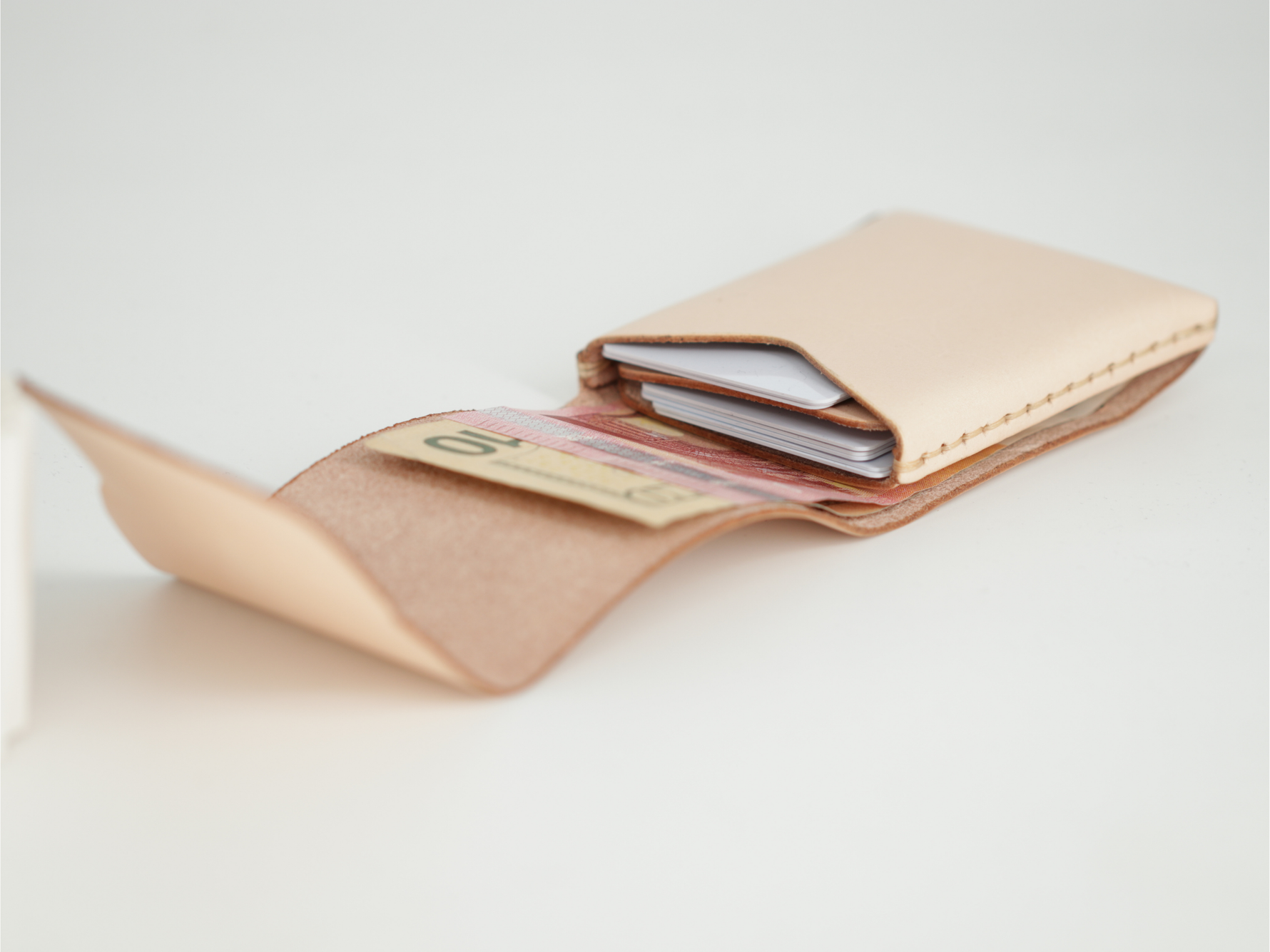A light beige leather card holder wallet shown open, containing credit cards and a banknote, on a clean white background. 