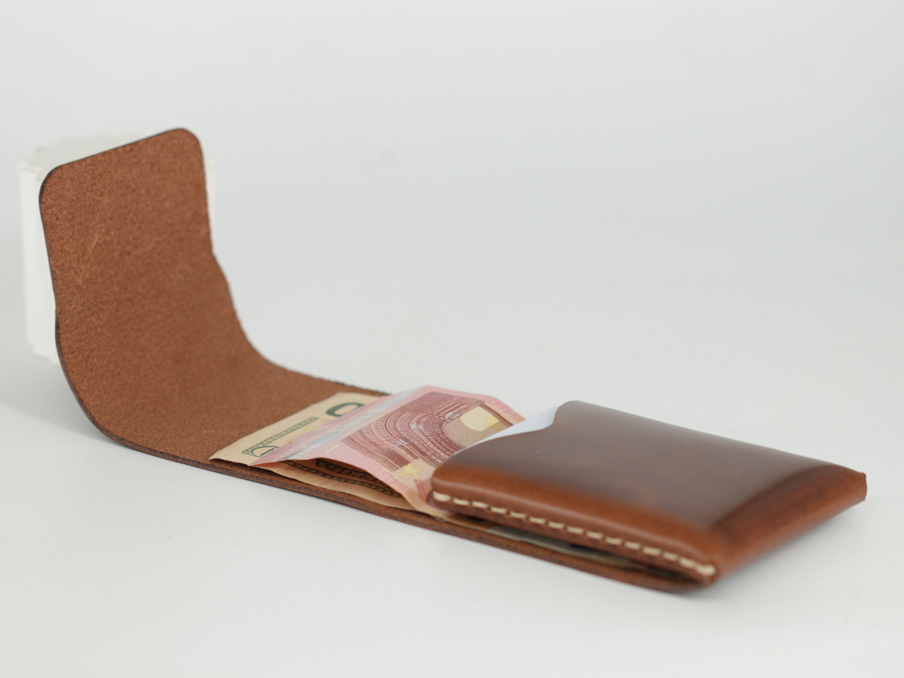 A brown leather card holder wallet shown open, displaying its interior lining and a pink banknote tucked inside, set against a plain light gray background. 