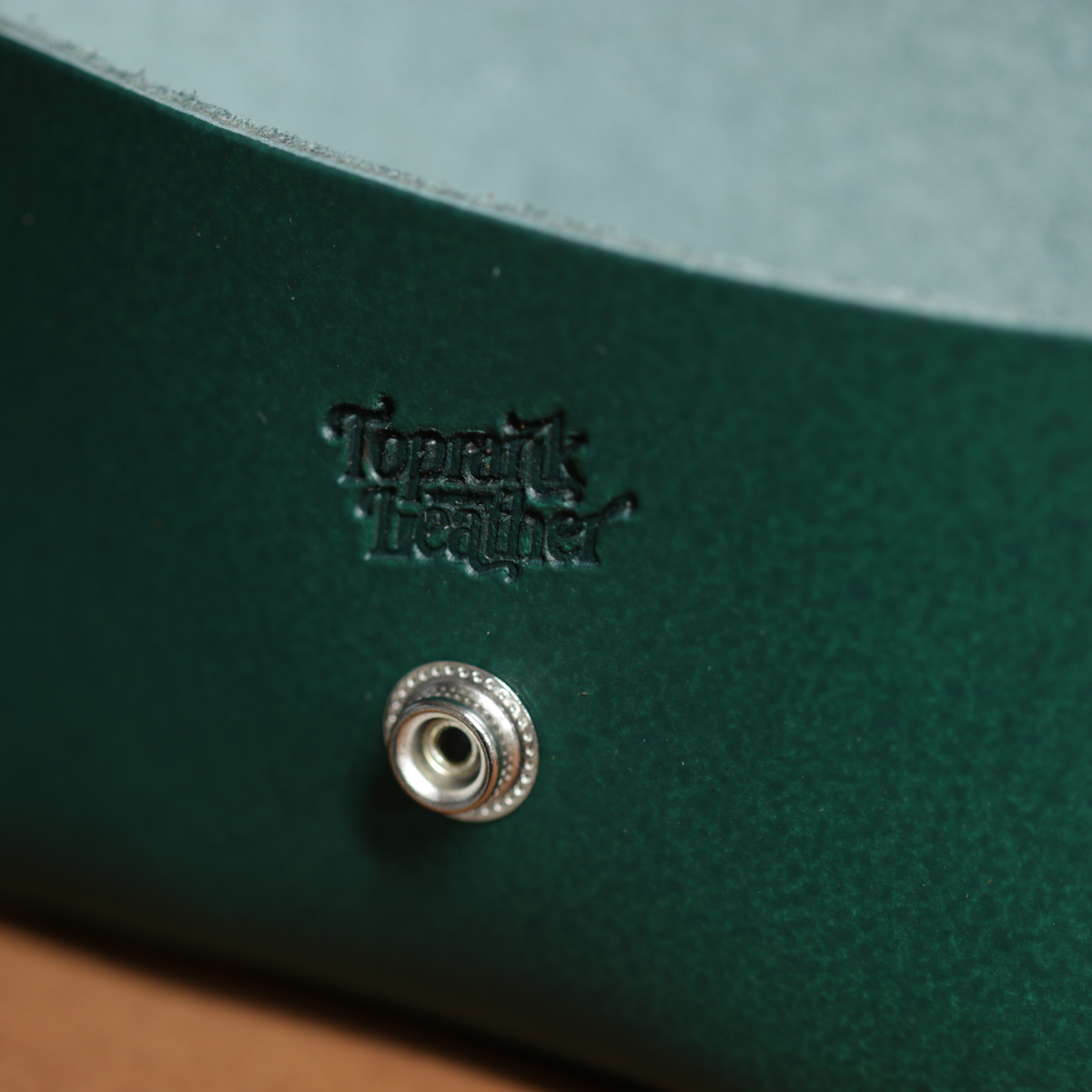 Close-up of a green leather product with a silver button and 'Toprank Leather' branding. 