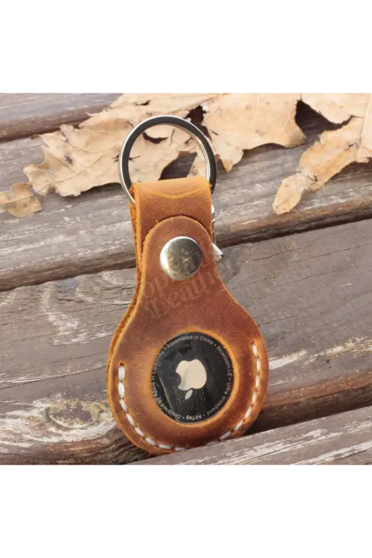 Brown leather keychain with a black circular device featuring an apple logo, on a wooden surface with leaves. 